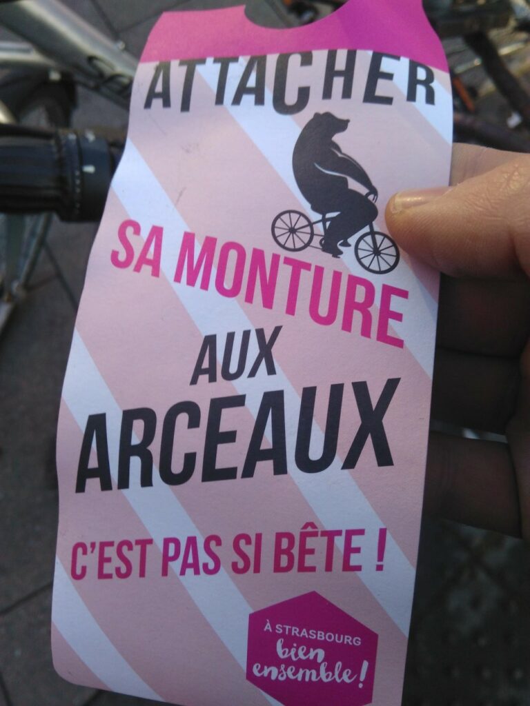 Un flyer type accroche porte avec écrit "Attacher sa monture aux arceaux c'est pas si bête" avec une image d'un ours sur un vélo