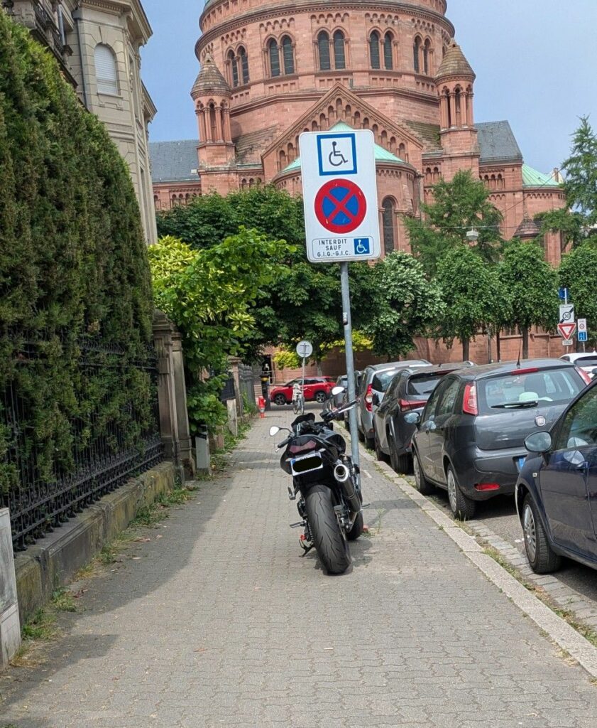 Une moto est stationnée au milieu du trottoir, attachée à un panneau d'interdiction de stationner, sauf pour les personnes en situation de handicap